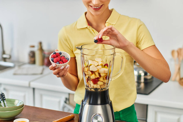 oříznutý pohled usměvavé ženy přidávající bobule do elektrického mixéru s nakrájeným ovocem, vegetariánská strava - Fotografie, Obrázek