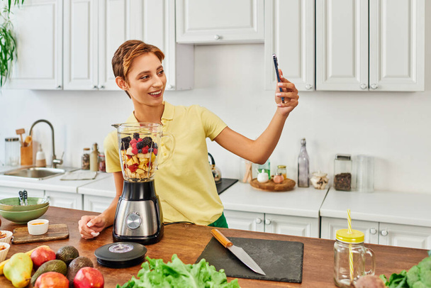 joyful vegetarian woman with smartphone taking selfie near blender with chopped fruits in kitchen - Foto, afbeelding