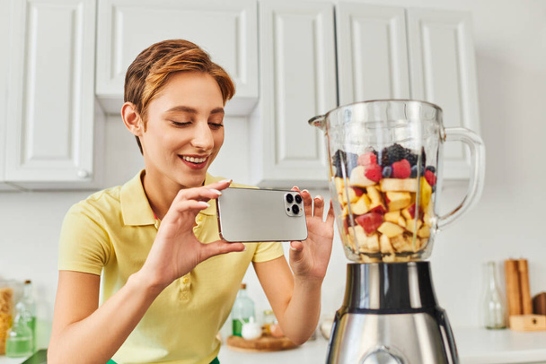 femme positive prenant des photos de mélangeur électrique avec des fruits hachés, délicieux régime à base de plantes - Photo, image
