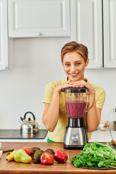 vegetarian woman smiling at camera near blender with wholesome smoothie in kitchen, plant-based diet - Photo, image