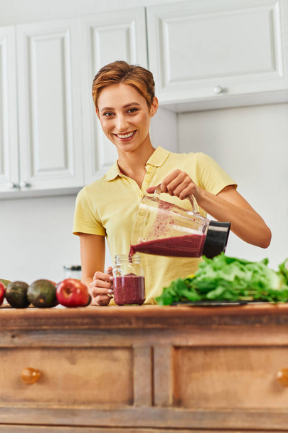vegetarian culinary delight, happy young woman pouring fresh smoothie from blender into mason jar - Photo, Image