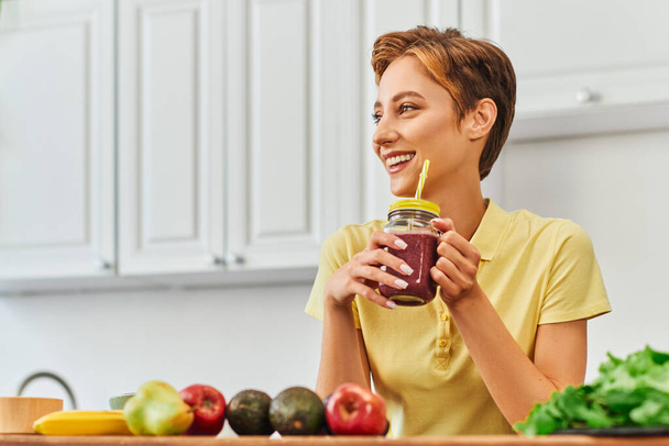 joyful vegetarian woman holding fresh smoothie in mason jar with straw and looking away in kitchen - Photo, Image