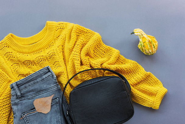 Vlakke lay van de gele trui van de vrouw, grijze jeans, zwarte tas en kleine pompoen, herfstblad op grijze achtergrond. Herfst modeconcept. Bovenaanzicht, vlak lay. - Foto, afbeelding