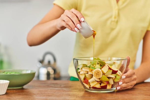 cropped view of woman pouring honey into bowl with chopped fruits, wholesome vegetarian salad - Photo, image