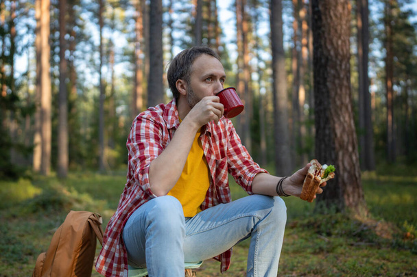Uomo escursionista bere il tè dalla tazza durante le escursioni nella foresta. Maschio durante la piccola pausa mangiare panino e bere caffè nei boschi autunnali, riposo fermato per una pausa dopo la passeggiata nei boschi.  - Foto, immagini