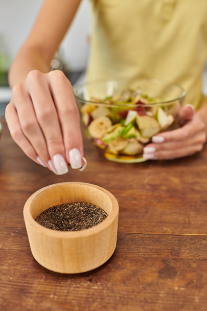oříznutá vegetariánka, která bere sezamová semínka z dřevěné mísy, zatímco doma připravuje ovocný salát - Fotografie, Obrázek