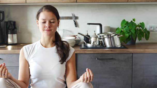 Genç bir kadın mutfakta oturmuş kirli bulaşıkların arka planı üzerine meditasyon yapıyor. Yavaşla, kendine dikkat et, yavaş yaşam konsepti. Yüksek kalite 4k görüntü - Video, Çekim