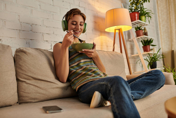 mujer alegre en auriculares teniendo merienda vegetariana por la noche en el sofá en la acogedora sala de estar - Foto, imagen