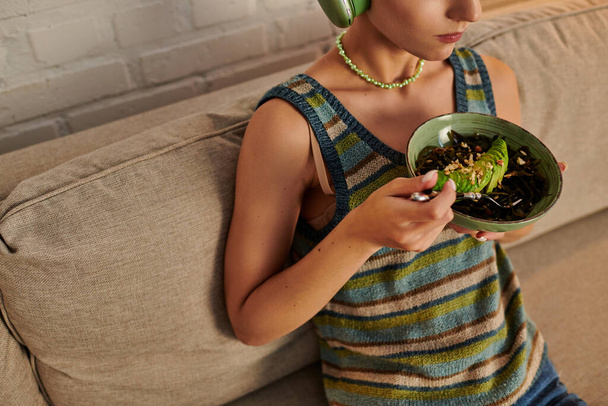 обрезанная вегетарианка сидит с тарелкой салата с морскими водорослями и нарезанным авокадо в гостиной - Фото, изображение