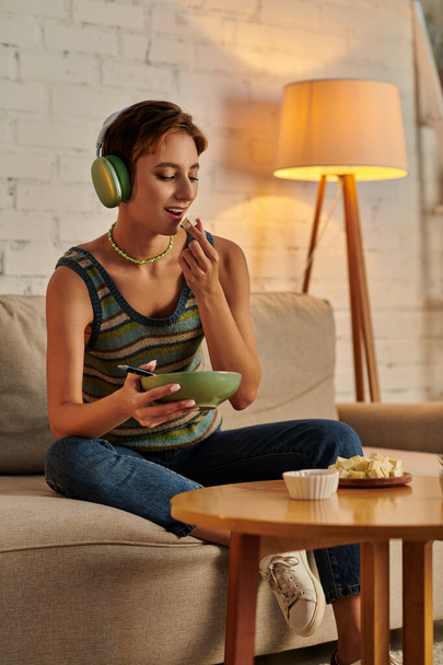 vegetarian woman in headphones holding salad bowl and eating tofu cheese, evening snack on sofa - Photo, Image
