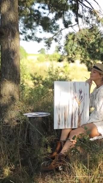 A girl artist paints a picture in a pine forest. Beautiful girl artist outdoors. Background landscape and creates creativity in the forest in summer. Vertical video. - Footage, Video