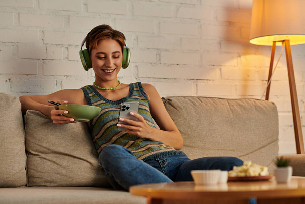 happy veggie woman in headphones networking on smartphone while sitting with salad bowl on couch - Photo, Image
