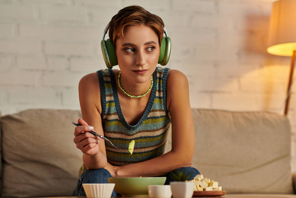 dreamy woman in headphones looking away while having evening vegetarian snack on couch at home - Photo, Image