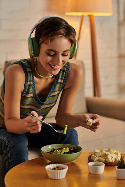 radostná žena ve sluchátkách večeří s vegetariánským salátem a tofu sýrem na pohovce doma - Fotografie, Obrázek