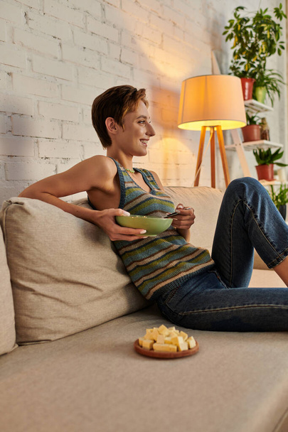 улыбающаяся женщина с салатной миской сидит на диване в гостиной рядом с сыром тофу, вегетарианский ужин - Фото, изображение
