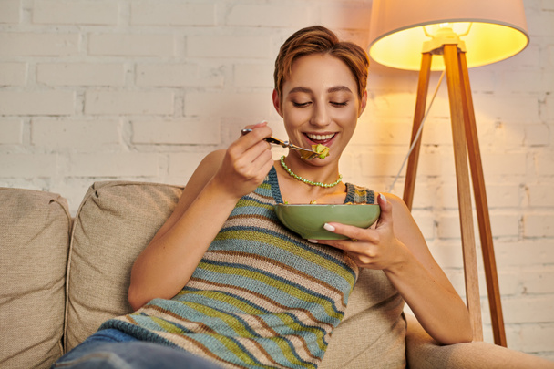 pleased young woman eating delicious vegetarian salad during evening snack in cozy living room - Photo, Image