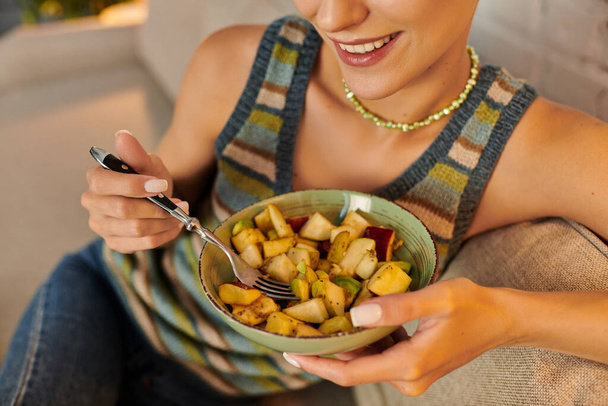 Evdeki kanepede bir kase lezzetli meyve salatasıyla gülümseyen kadın manzarası, bitki temelli diyet. - Fotoğraf, Görsel