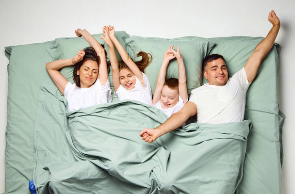 Вид сверху на симпатичных симпатичных привлекательных людей, накрытых одеялом, лежащих в кровати, поднимающих в пробуждении распростертые руки. Семья в одной постели спит. Концепция хорошего сна, семейное время, бодрствование - Фото, изображение