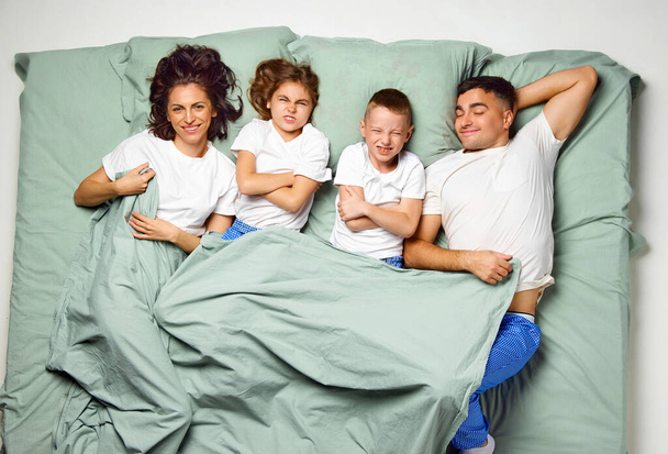 Vista dall'alto di persone coperte di coperta sdraiate a letto. Bambini mani incrociate e genitori felici e simpatici. Famiglia in un letto sveglio. Concetto di benessere del sonno, tempo in famiglia, sveglio, amore, mattina - Foto, immagini