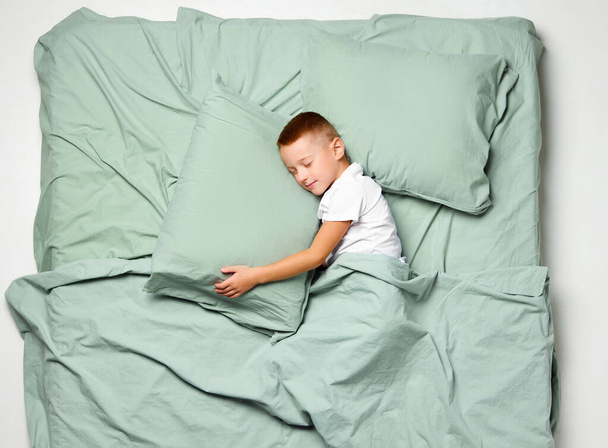 Retrato de vista superior do menino deitado de lado coberto com cobertor dormindo e abraçando travesseiro. Miúdo perturbado, criança em grande cama de menta. Conceito de emoções, bem-estar do sono, toque humano, soneca, hora de dormir. Anúncio - Foto, Imagem