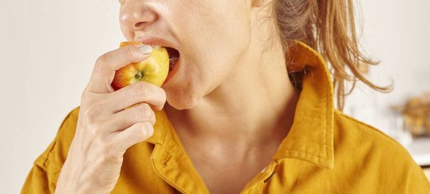 Woman eating apple, close-up, hand and mouth. Kitchen background. - Photo, Image