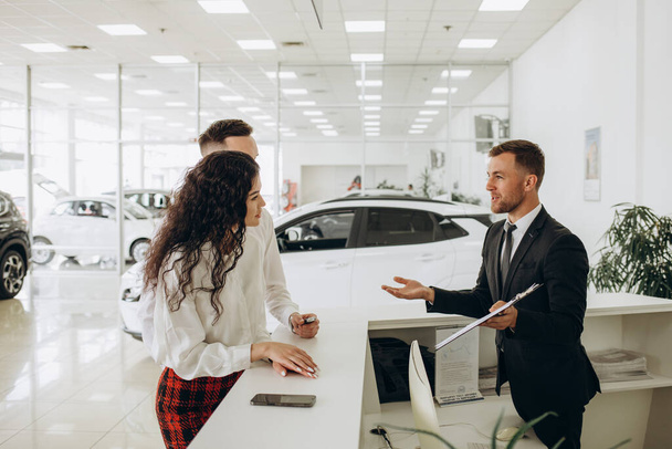 Verkaufsleiter zeigt einem jungen Paar eine Auto-Ladestation und verkauft im Showroom Elektroautos. Konzept des Kaufs umweltfreundlicher Autos für die Familie. Hochwertiges Foto - Foto, Bild