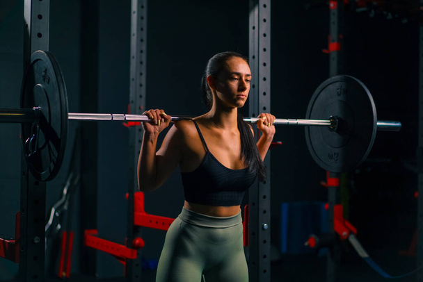 retrato de joven hermosa mujer de fitness enfocada haciendo ejercicio de presión de hombro pesado con barra gimnasio entrenamiento grupos musculares - Foto, Imagen