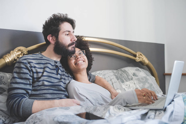 young multiethnic couple indoor using computer sitting bed  - Photo, Image