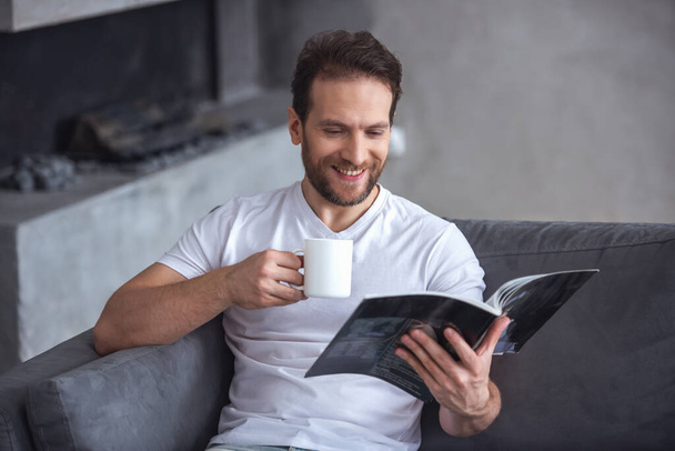 Knappe man is lezen van een tijdschrift, een kopje holding en glimlachen terwijl rusten thuis - Foto, afbeelding