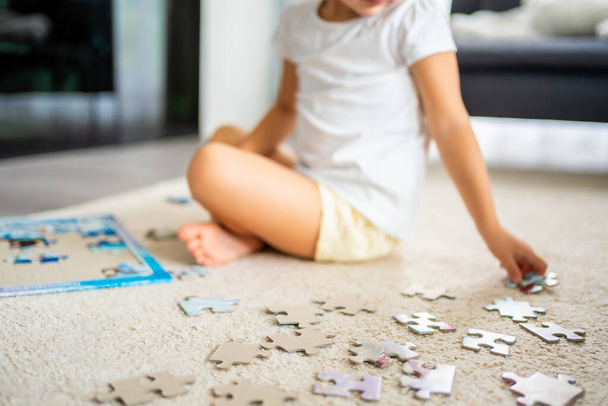 Little blonde girl sits at home on the carpet and collects puzzles. Focus on the puzzle pieces. High quality photo - Photo, Image