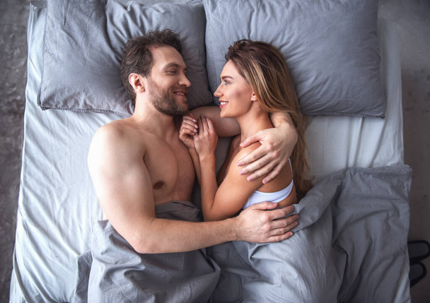Вид сверху красивой пары, обнимающейся и улыбающейся, лежа вместе в постели дома
 - Фото, изображение