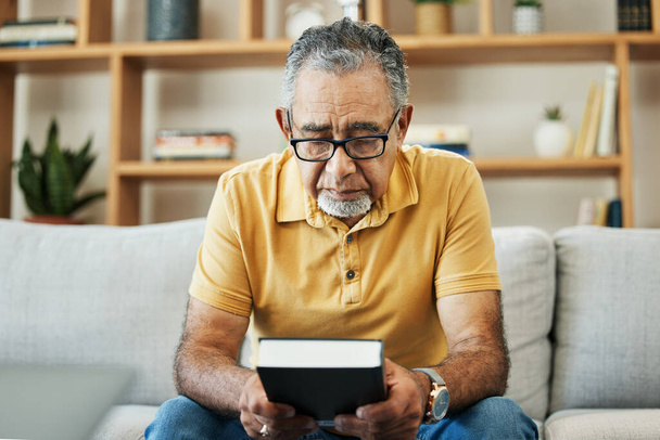 Mature, man and bible by thinking in home, alone and sadness of memory in living room of thoughts. Elderly, person or grandfather on sofa with hand, holding and book for religion, church or practice. - Photo, Image