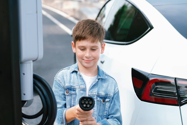 環境に優しいEV車と小さな男の子がカメラでEV充電器を指している道路旅行の休暇. 環境保護のためのクリーンで持続可能なエネルギーを使用して電気自動車および充電ステーション. パーペチュアル - 写真・画像