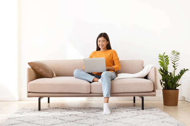Glückliche attraktive junge koreanische Frau digitalen Nomaden mit Laptop zu Hause, sitzt auf der Couch im gemütlichen Wohnzimmer, Kopierraum. Positive asiatische Dame, die aus der Ferne arbeitet, mit Freunden chattet, online einkauft - Foto, Bild