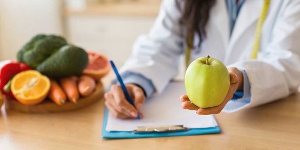 Kobieta lekarz na receptę lub wypisywanie planu diety i trzymanie zielonego jabłka, siedzi przy biurku ze świeżymi owocami, przycięte, panorama, zbliżenie - Zdjęcie, obraz