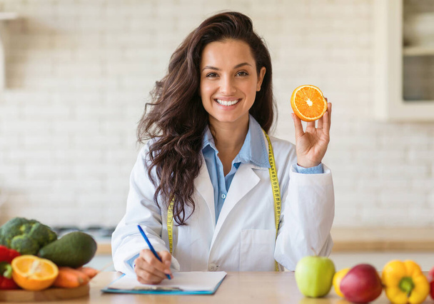Attractive young european nutrition adviser holding orange and making meal plan for client, working at weight loss clinic. Smiling female dietitian writing prescription for patient - Photo, Image