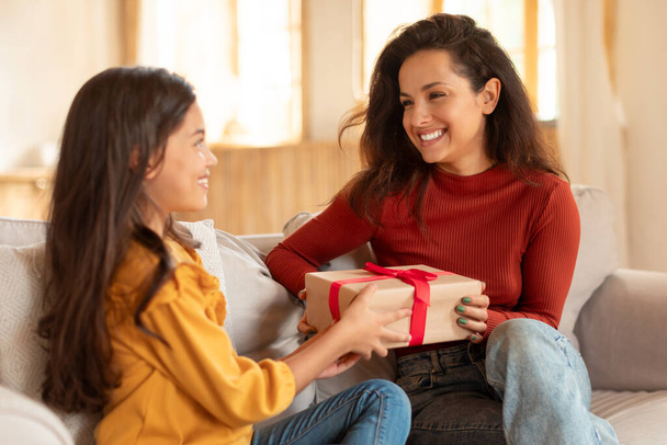 Happy Daughter Surprising Mother Giving Birthday Wrapped Gift To Her Sitting On Couch Indoor, Celebrating Moms Day Holiday Together, Kid Presenting Box To Cheerful Mommy At Home - Photo, Image