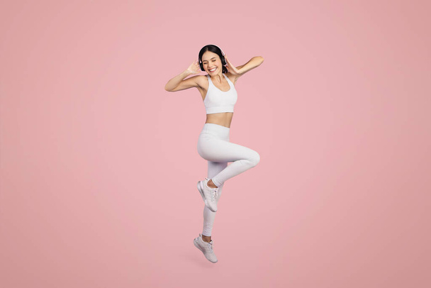 Mujer excitada en ropa deportiva y auriculares inalámbricos saltando sobre el fondo rosa, disfrutando de música favorita durante el entrenamiento, tiro de cuerpo entero, espacio libre - Foto, imagen