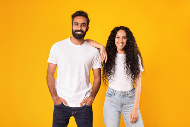 Studioaufnahme eines glücklichen romantischen arabischen Paares, das zusammen steht, eine Frau, die sich auf einen Mann stützt, während sie über gelbem Hintergrund posiert. Junge Ehepartner in lässigen weißen T-Shirts lächeln in die Kamera - Foto, Bild