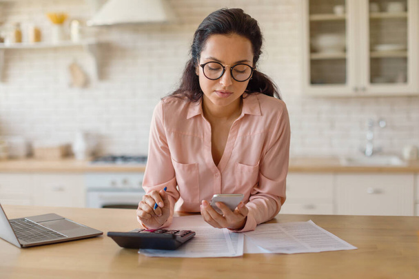 Young lady with cellphone and calculator accounting taxes and planning budget, sitting at table in kitchen interior, woman calculating spends and checking financial documents - 写真・画像