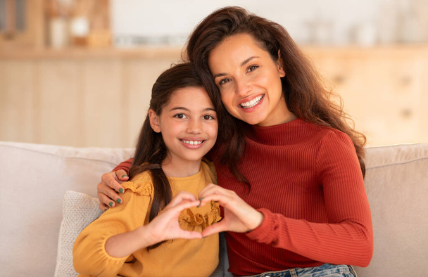 Χαμογελαστή μητέρα και κόρη που δείχνουν το σύμβολο της καρδιάς με τα δάχτυλα στην κάμερα, ενώνοντας τα χέρια τους μαζί σαν σύμβολο καλοσύνης και αγάπης, καθισμένοι στον καναπέ στο σπίτι. Δεσμός παιδιού και μαμάς - Φωτογραφία, εικόνα