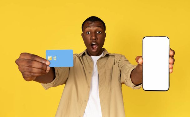Järkyttynyt iloinen tuhatvuotinen afrikkalainen amerikkalainen kaveri näyttää puhelin tyhjä näyttö ja luottokortti, eristetty keltainen studio tausta. Mainos ja tarjous, sovellussuositus, pankkitoiminta ja myynti, verkkokauppa - Valokuva, kuva