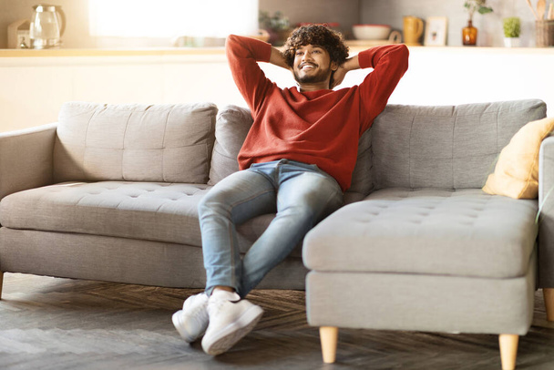 Domestic Comfort. Happy Young Indian Man Relaxing On Comfortable Couch In Living Room, Smiling Millennial Eastern Man Leaning Back With Hands Behind Head, Enjoying Weekend Leisure, Copy Space - Photo, Image