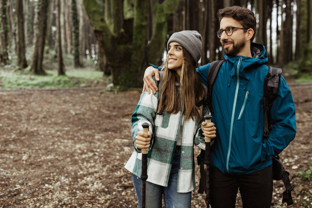 Glad young caucasian guy hug lady in jackets with trekking sticks enjoy adventure, spare time, walk in cold forest in autumn. Modern vacation together, active lifestyle at weekend - Photo, Image