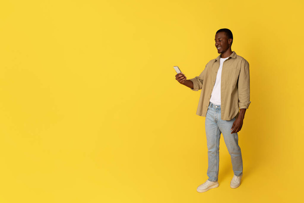 Smiling millennial african american man in casual use phone for chat, isolated on yellow studio background, full length. Communication remotely app, social networks, blog, good news - Photo, Image
