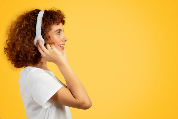 Happy teenager caucasian curly girl in wireless headphones listening music, enjoy free time, isolated on orange studio background, profile. Lifestyle, rest, relax, ad and offer - Foto, Imagem