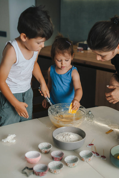 Una pequeña linda hija e hijo ayudando a su madre a preparar la masa para hornear pastel en la cocina en casa, concepto de familia feliz - Foto, imagen