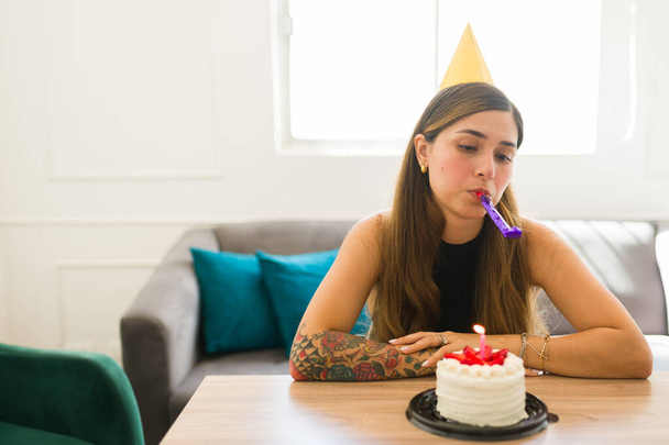 Depresyondaki Latin kadın evde tek başına doğum gününü kutlamak ve pasta yemek için parti ıslığı çalarken üzgün hissediyordu. - Fotoğraf, Görsel
