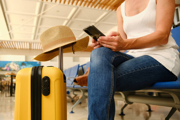 Crop donna anonima in abiti casual seduto sulla sedia vicino a valigia gialla e smartphone di navigazione in attesa di volo - Foto, immagini