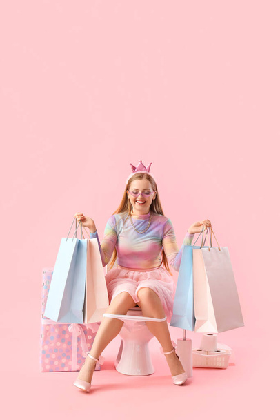 Young woman with shopping bags sitting on toilet bowl against pink background - Photo, Image